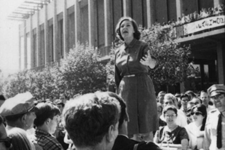 ID: grayscale image of Jackie Goldberg standing on a car and speaking to a crowd; behind her is the east side of thein front of the Martin Luther King Jr. Student Union