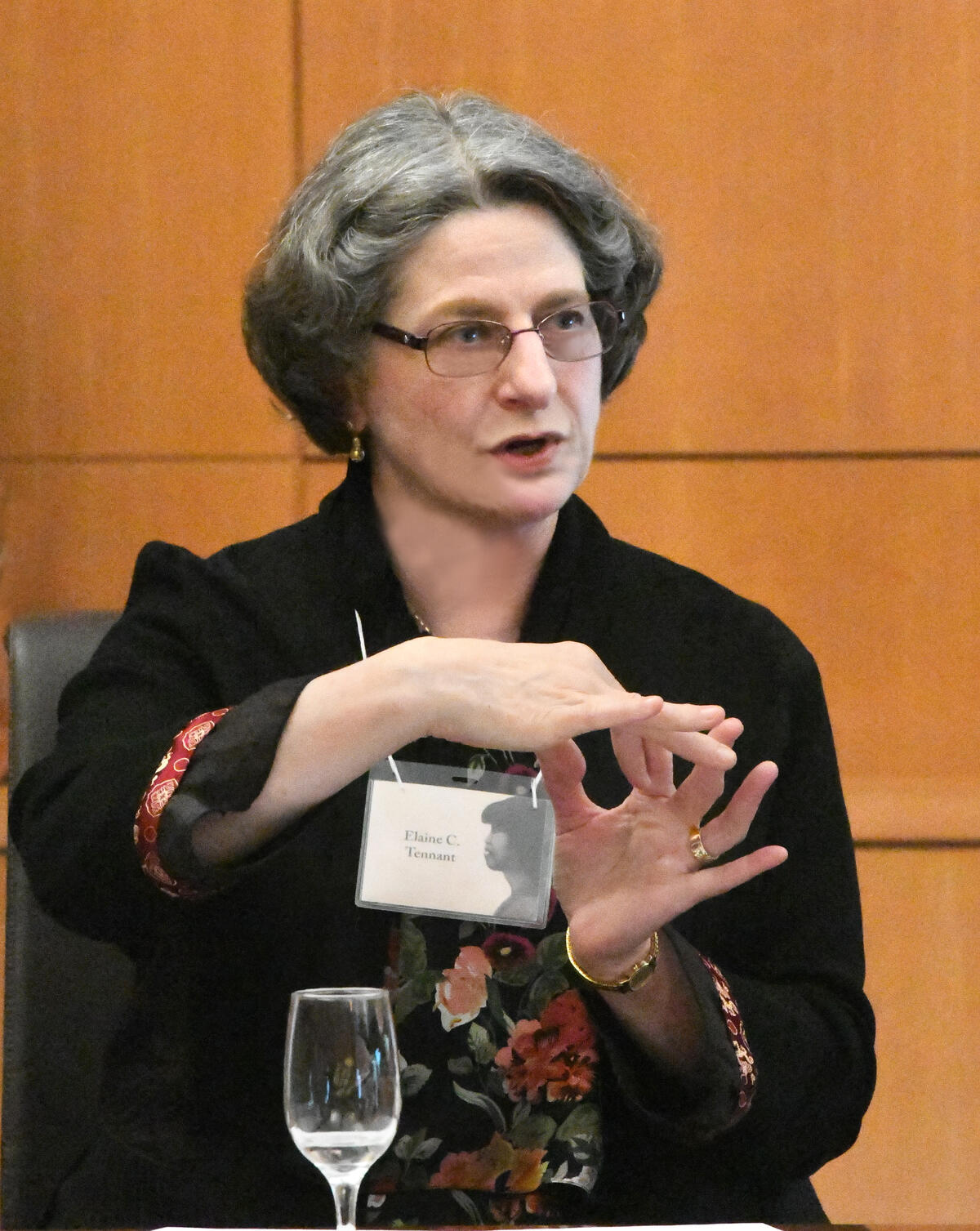 ID: image of Elaine Tennant mid-speech in glasses,  floral black top, name tag, and jewelry using hand gestures to illustrate a point