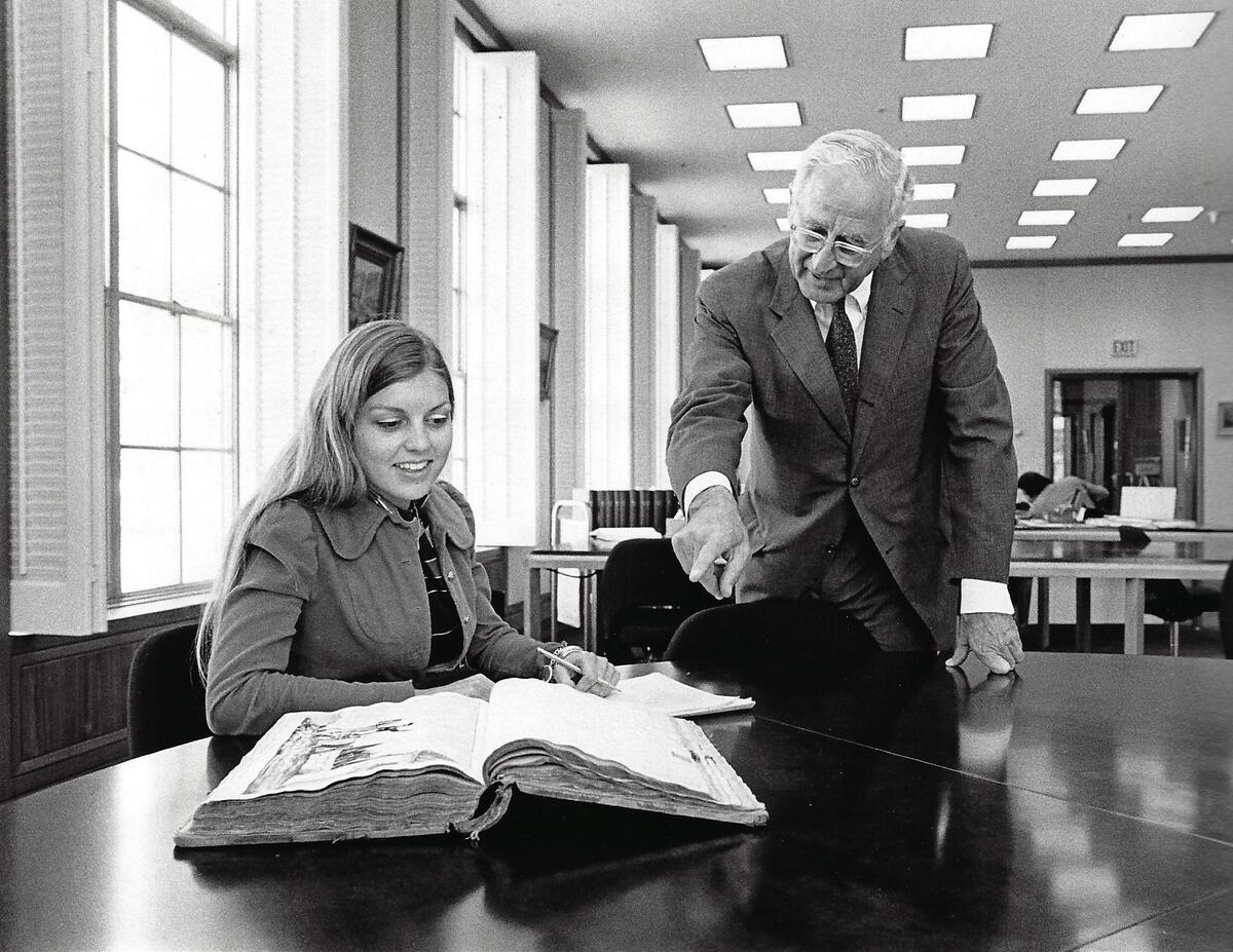ID: Suzanne Calpestri smiling and taking notes in the Bancroft library, a man in a suit pointing.