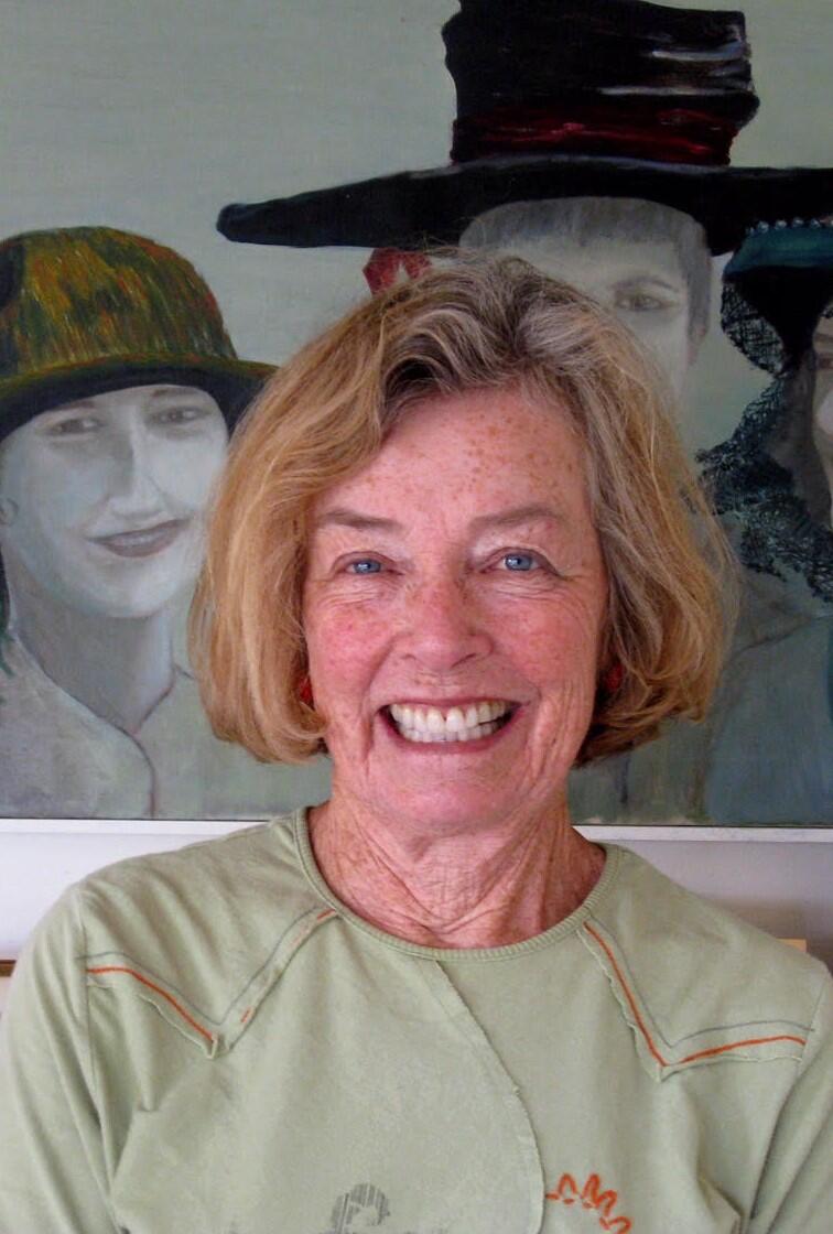 ID: Suzanne Reiss smiling in short haircut and green shirt in front of a painting of 3 women