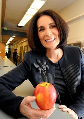  Barbara Davis in black cardigan adorned with silver floral brooch, holding an apple in one hand and smiling directly into camera