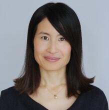  headshot of Carolyn Chen smiling with hair flipped out in necklace and dark top against gray background 