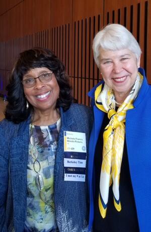  Michele Woods-Jones smiling into camera, wearing a blue blazer over blue and green patterned blouse with nametag on the left side. Carol Christ smiles into camera with royal blue blazer over black blouse with yellow patterned scarf