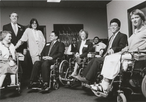  grayscale photo of 8 people using wheelchairs dressed in formal attire as DSRP participants; there are 2 people standing and smiling into the camera; in the far left is Susan O'Hara in candid conversation with an unidentified man