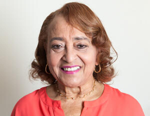  headshot of Jewelle Taylor Gibbs, smiling towards the camera and wearing a salmon colored blouse, golden hoop earrings, and gold accented necklace