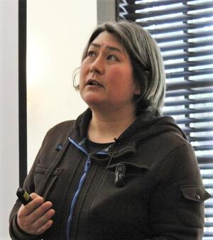  candid photo of Professor Karen Nakamura mid-speech giving a presentation, holding a slideshow clicker, wearing a black jacket