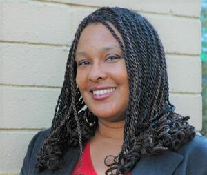  Michele de Coteau, PhD in black blazer and red blouse, looking into the camera at a 45 degree angle, against a gray brick wall