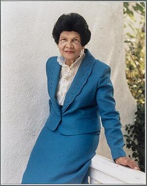  photo of Miriam Matthews smiling in red lipstick and wearing a dark cerulean blue suit and white lace blouse, posed against a cream colored wall