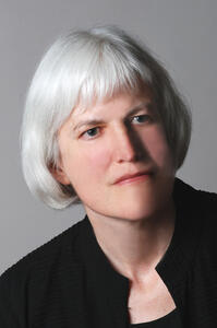  headshot of Georgina Kleege with neutral expression and white bob haircut with bangs in black top against a gray background
