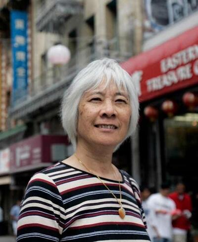  photo of Judy Yung in short white hair and gold necklace in striped shirt, smiling at the camera; behind her is a blurred background of SF Chinatown