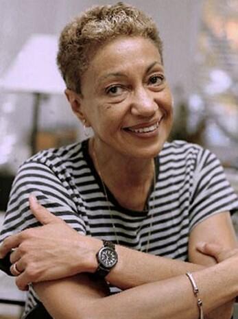  June Jordan smiling into the camera with head slightly tilted, arms crossed in relaxed manner, wearing a black and white striped t-shirt and black watch
